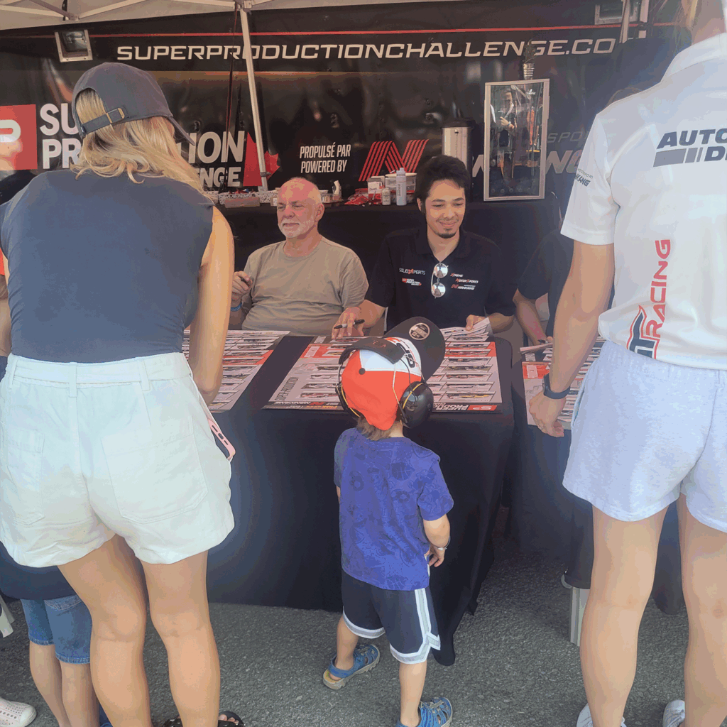Racing driver Michael Habrich signs posters for a young boy wearing ear protection, with other fans gathered around the autograph tent.