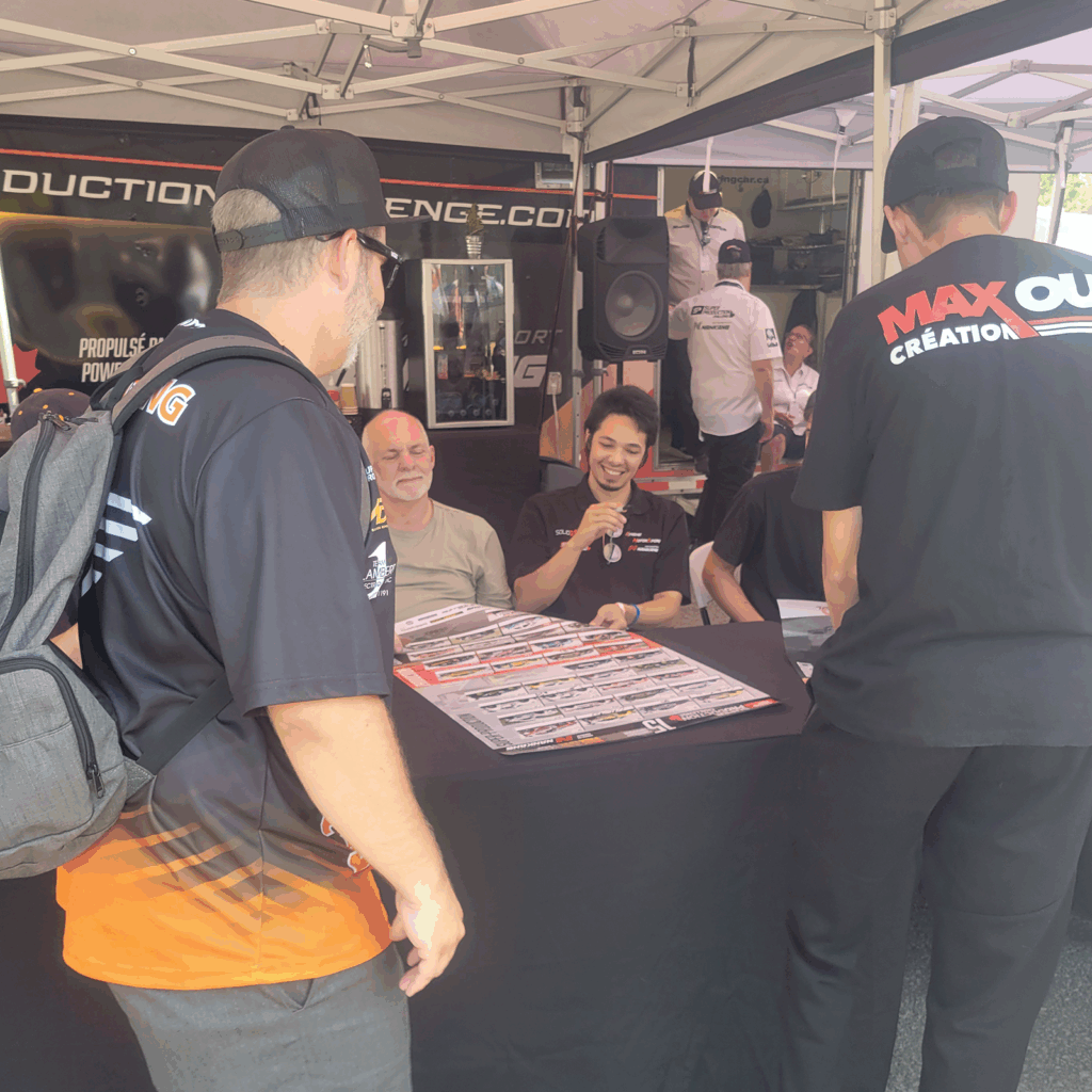 Racing drivers sit at an autograph table under a canopy during GP3R, smiling and interacting with fans standing in line.