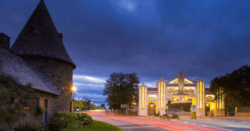 Scenic view of the illuminated Trois-Rivières city entrance arch at twilight, with streaks of car lights on the road and a stone tower on the left.