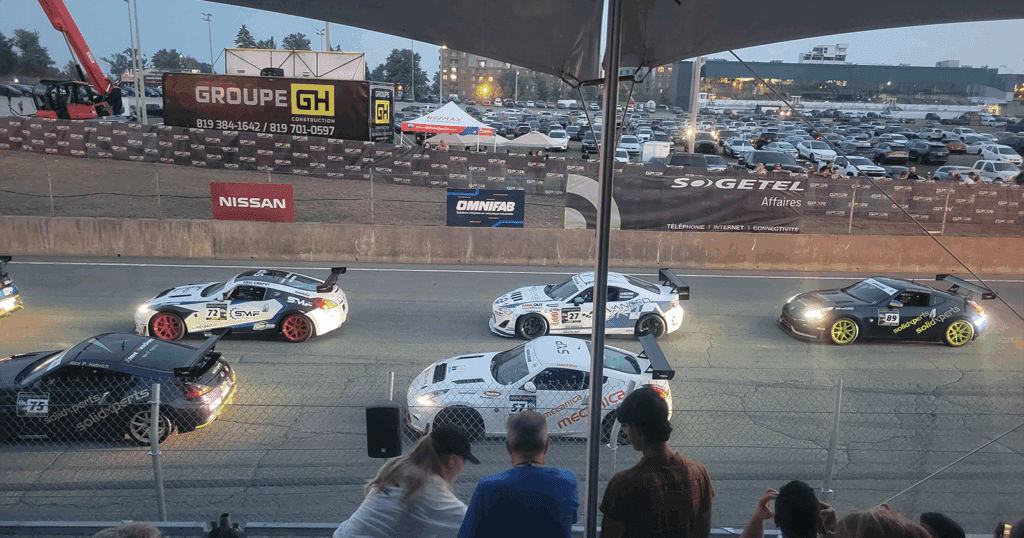 Evening shot from a shaded grandstand of multiple race cars lined up on the starting grid at GP3R, with spectators watching from the foreground.