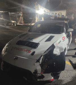 White Mecanica Nissan 370Z race car with visible front-end damage parked in the paddock under bright lights after the race.