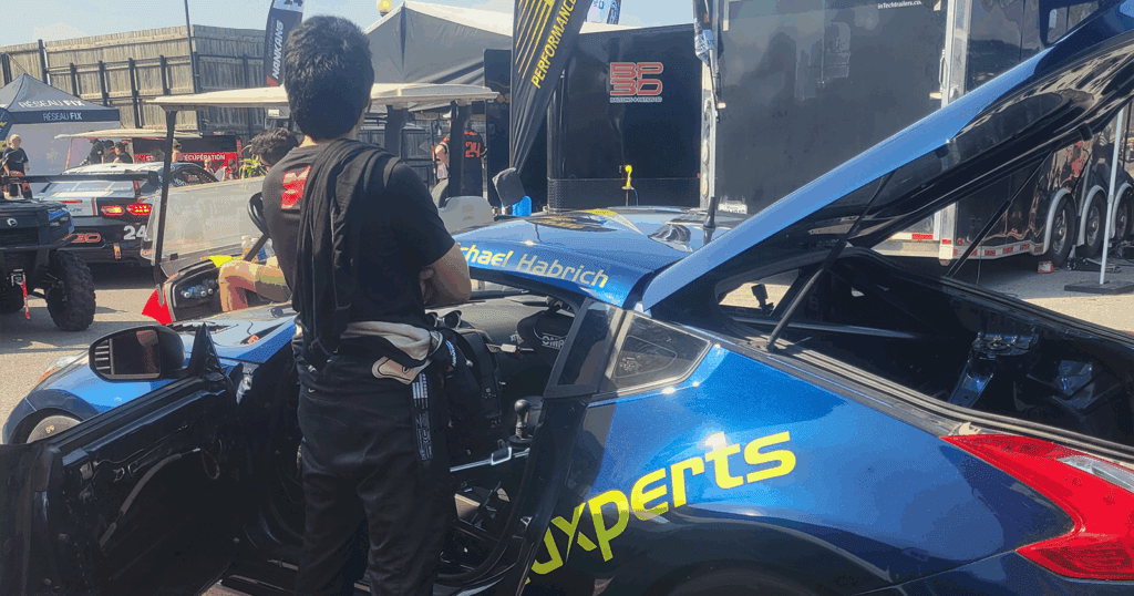 A driver in a racing suit prepares inside a dark blue Solidxperts Nissan 370Z with neon green wheels in the paddock area during the day.