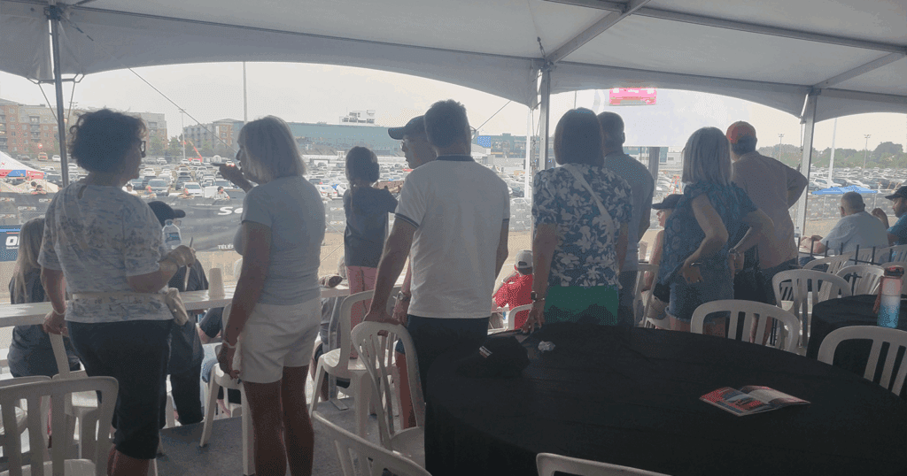 Group of spectators standing under a tented corporate lounge, looking out toward the racetrack with cars passing in the background.