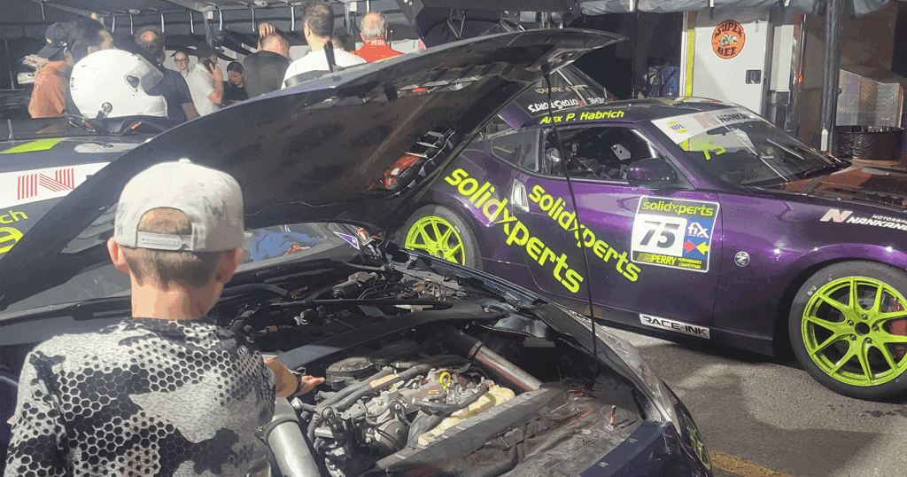 A boy looks at the engine bay of a blue Solidxperts Nissan 370Z while a purple Solidxperts 370Z is parked nearby in the paddock at night.
