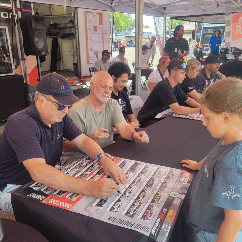 Racing driver Alex Habrich signs posters at a long autograph table while fans, including a young girl, line up during the Grand Prix 3R event.